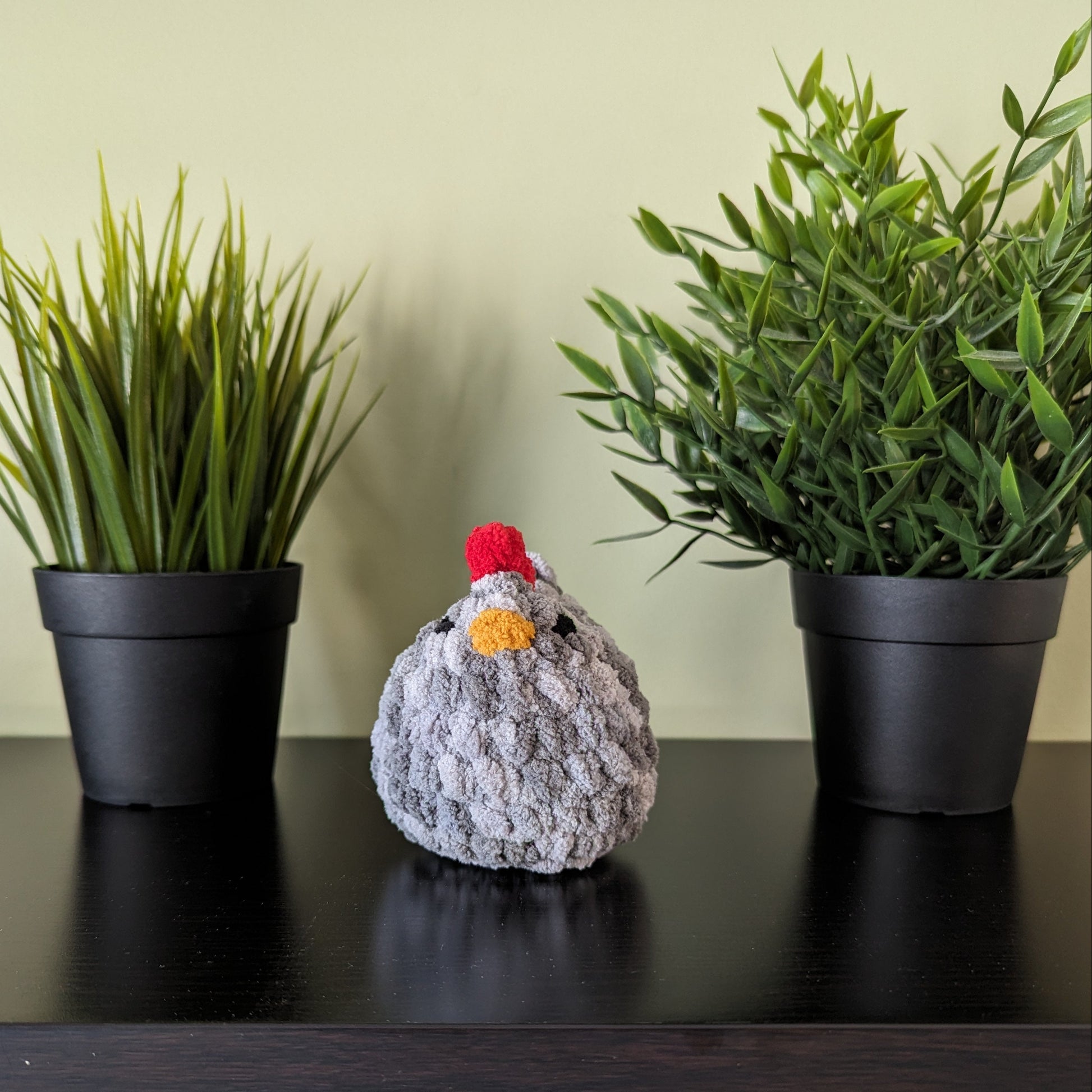 A grey plush crochet chicken with a red comb and yellow beak. The chicken is sitting on a wooden table. There is a green plant in a white pot in the background. There are shelves above the table with jars and containers.