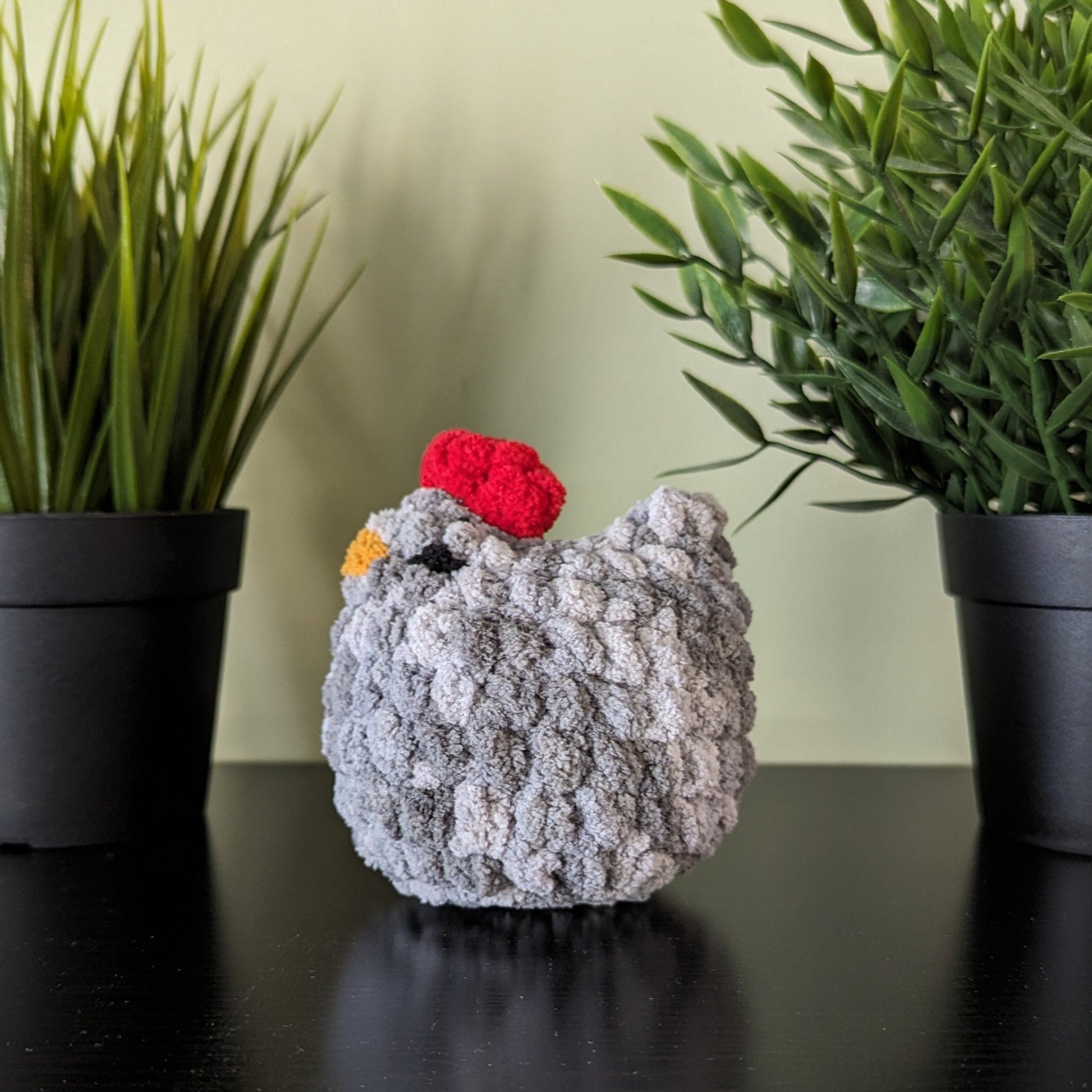 A plush grey crochet chicken with a red comb and yellow beak. The chicken is sitting on a black shelf. The wall in the background is pale green. There are two artificial green plants in black pots on either side of the chicken on the shelf.